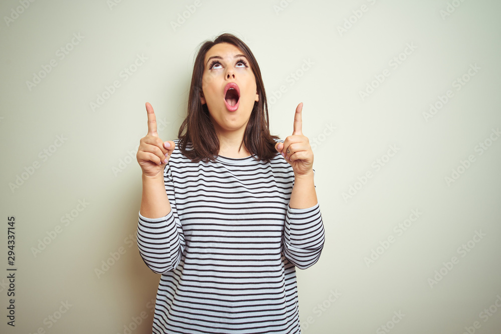 Young beautiful brunette woman wearing striped sweater over white isolated background amazed and surprised looking up and pointing with fingers and raised arms. Young beautiful brunette woman wearing striped sweater over white isolated background amazed and surprised looking up and pointing with fingers and raised arms.