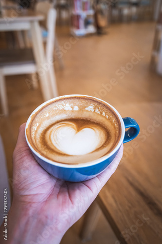 Man holding blue cup of hot cappuccino coffee with latte art on wooden coffee table, selective focus for drink background