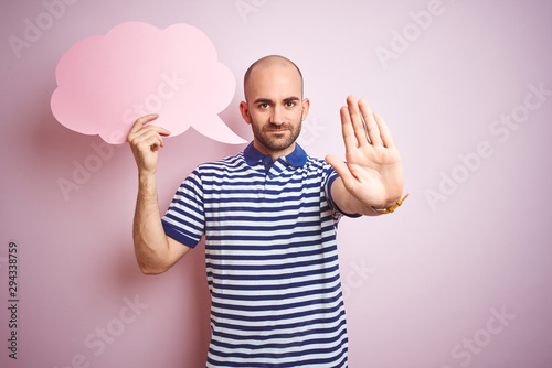 Young man holding speech bubble with blank space for message over pink isolated background with open hand doing stop sign with serious and confident expression, defense gesture
