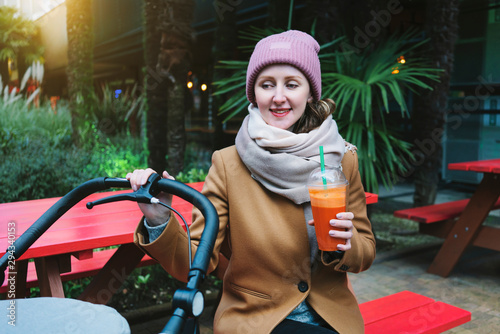 Portrait of young woman sitting at open-air food court with stroller. Outdoor terrace for visitors with children and baby vehicles in city cafe. Comfortable recreation area to have drink or order food