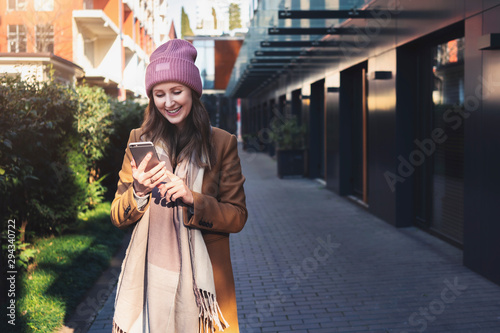 Girl uses smartphone while walking in urban district with modern architecture. Female wearing coat, cap and using smartphone app.