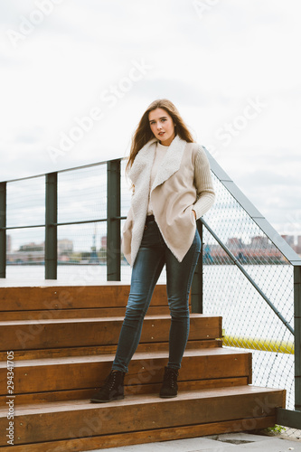 Beautiful young long-haired Caucasian woman stands on steps of wooden platform on waterfront at pier in port.