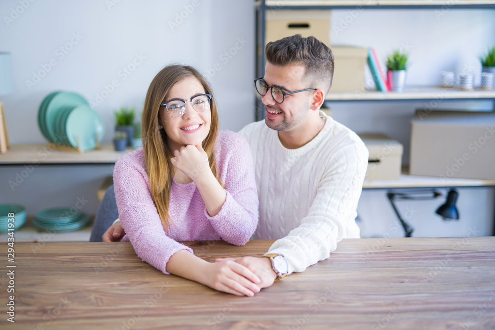 Young beautiful couple sitting on the table at home, hugging in love very happy for moving to new home with cardboard boxes behind them Young beautiful couple sitting on the table at home, hugging in love very happy for moving to new home with cardboard boxes behind them