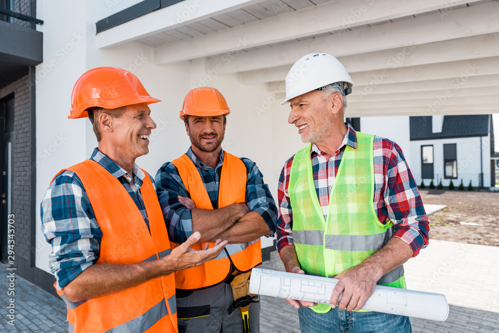 happy constructors in helmets standing near coworker with blueprint happy constructors in helmets standing near coworker with blueprint