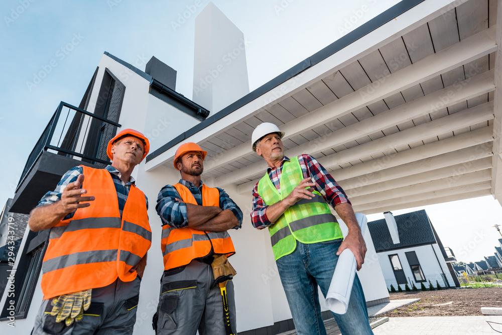 low angle view of constructors in helmets standing near coworker with blueprint low angle view of constructors in helmets standing near coworker with blueprint