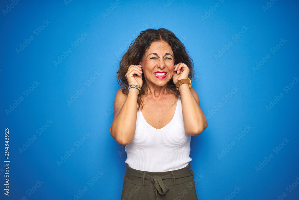 Middle age senior woman with curly hair standing over blue isolated background covering ears with fingers with annoyed expression for the noise of loud music. Deaf concept. Middle age senior woman with curly hair standing over blue isolated background covering ears with fingers with annoyed expression for the noise of loud music. Deaf concept.