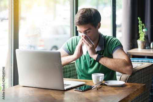 Young cold sick businessman in green t-shirt sitting at work, sneezing and cleaning his nose with tissue napkin. business and helthcare concept. indoor shot near big window at daytime.
