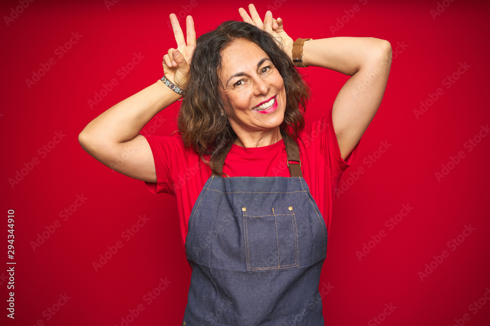 Middle age senior woman wearing apron uniform over red isolated background Posing funny and crazy with fingers on head as bunny ears, smiling cheerful Middle age senior woman wearing apron uniform over red isolated background Posing funny and crazy with fingers on head as bunny ears, smiling cheerful