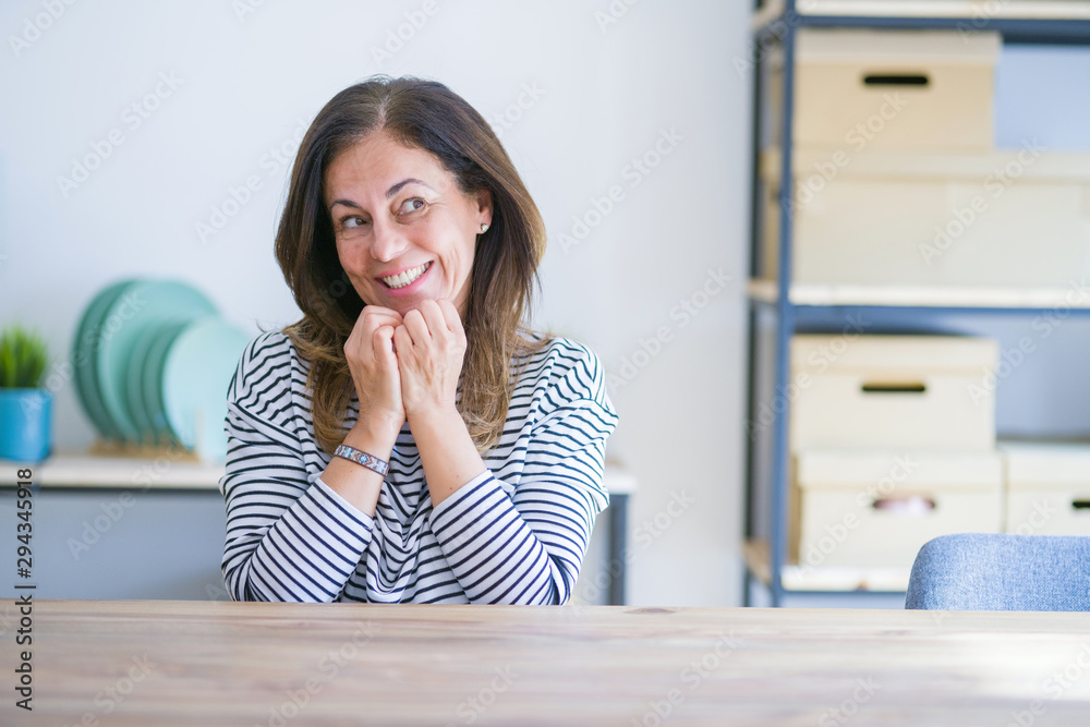 Middle age senior woman sitting at the table at home laughing nervous and excited with hands on chin looking to the side Middle age senior woman sitting at the table at home laughing nervous and excited with hands on chin looking to the side