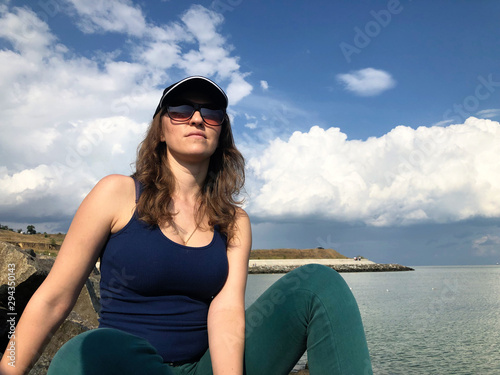 Girl on the beach in a baseball cap and sunglasses, on a background of blue sky and white clouds.