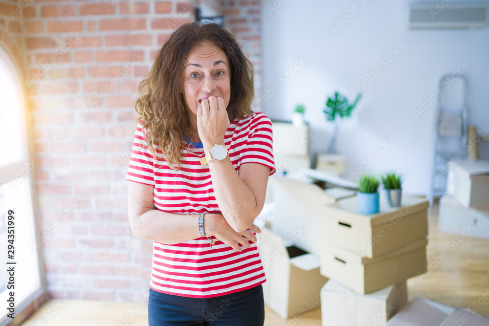 Middle age senior woman moving to a new house packing cardboard boxes looking stressed and nervous with hands on mouth biting nails. Anxiety problem.
