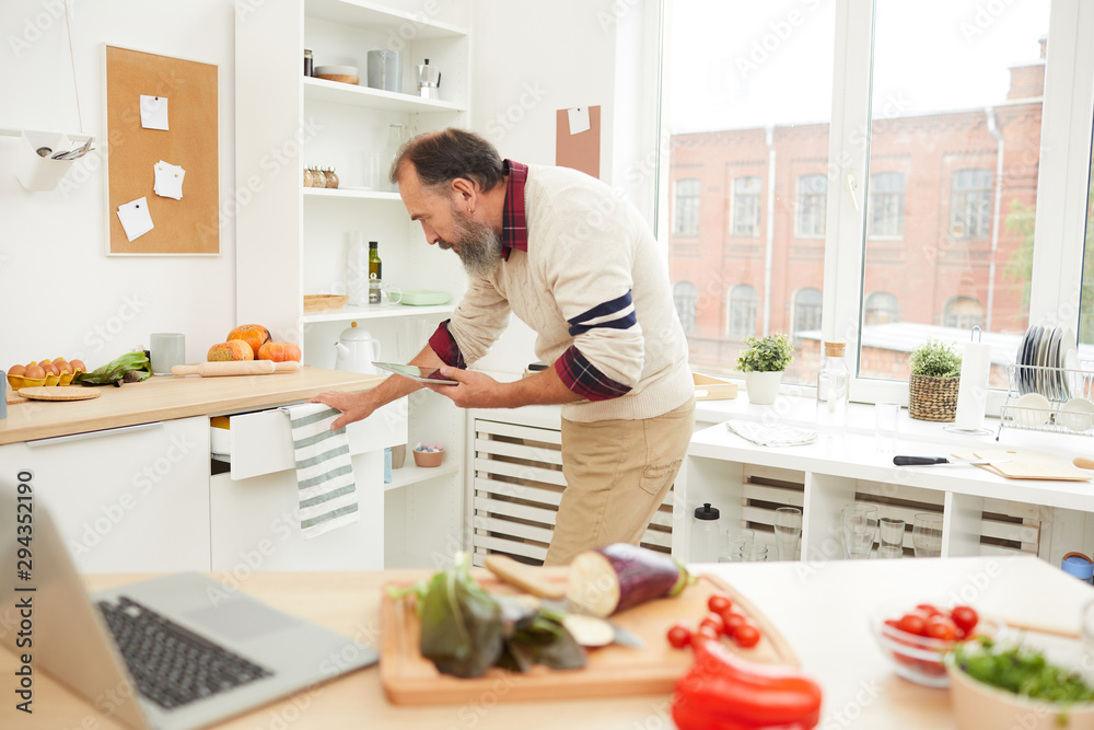 Side view portrait of bearded senior man opening drawers while cooking in kitchen, copy space Side view portrait of bearded senior man opening drawers while cooking in kitchen, copy space