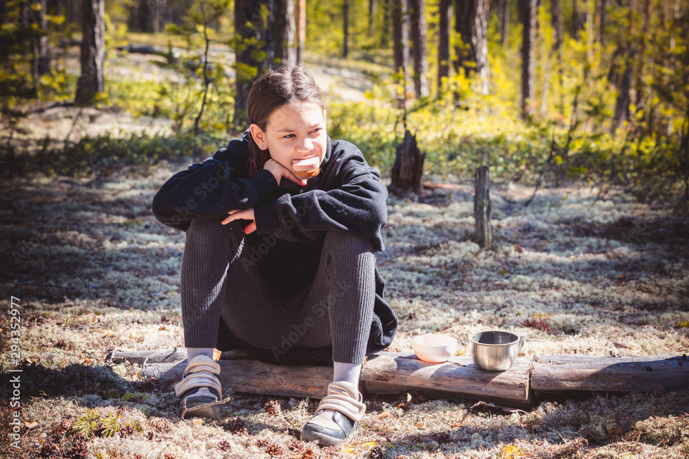 Fototapeta premium An adult girl in warm sweater sits on fallen tree and eats sandwich with sausage resting after hike in pine forest.
