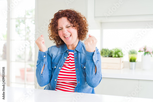 Middle age senior woman with curly hair wearing denim jacket at home very happy and excited doing winner gesture with arms raised  smiling and screaming for success. Celebration concept.