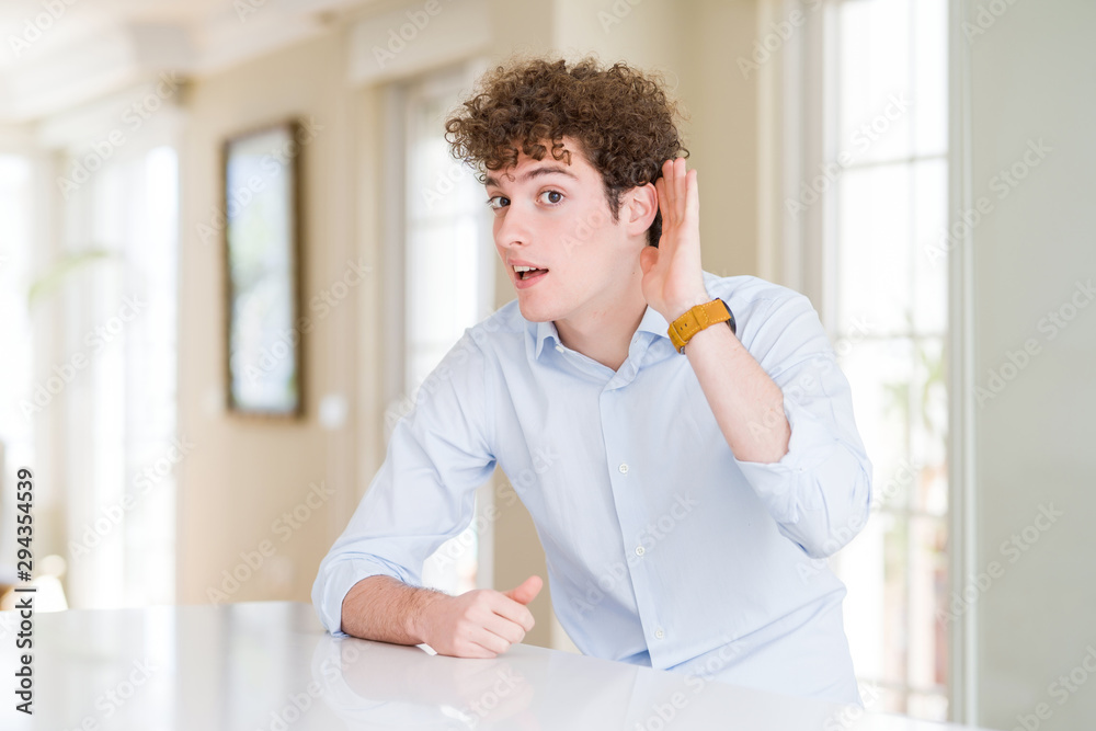 Young business man with curly read head smiling with hand over ear listening an hearing to rumor or gossip. Deafness concept.