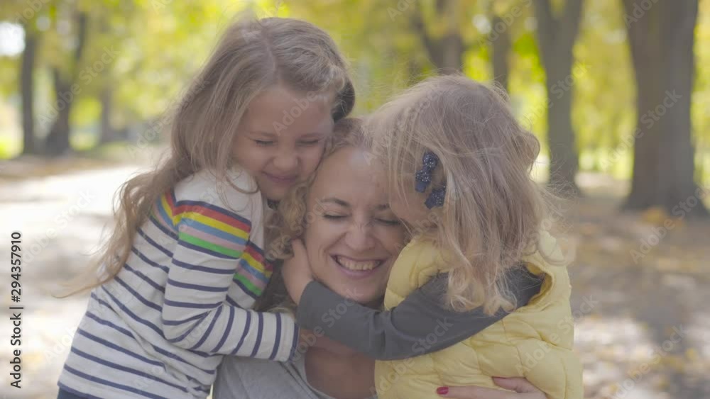 Close-up portrait of two joyful caucasian little sisters hugging curly blond woman in the autumn park. Cute smiling children expressing love to their adorable mother. Close-up portrait of two joyful caucasian little sisters hugging curly blond woman in the autumn park. Cute smiling children expressing love to their adorable mother.