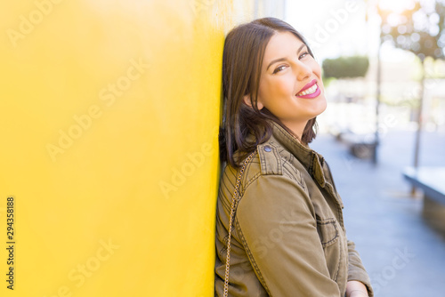 Beautiful young woman smiling confident and cheerful leaning on yellow wall, walking on the street of the city on a sunny day