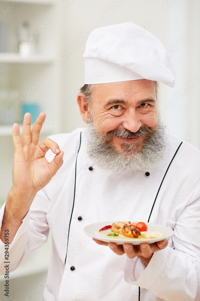 Portrait of cheerful senior chef presenting beautiful Italian dish and showing OK sign while posing in restaurant kitchen Portrait of cheerful senior chef presenting beautiful Italian dish and showing OK sign while posing in restaurant kitchen