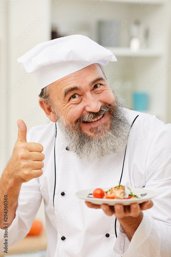 Portrait of cheerful senior chef presenting beautiful Italian dish and showing thumbs up while posing in restaurant kitchen Portrait of cheerful senior chef presenting beautiful Italian dish and showing thumbs up while posing in restaurant kitchen