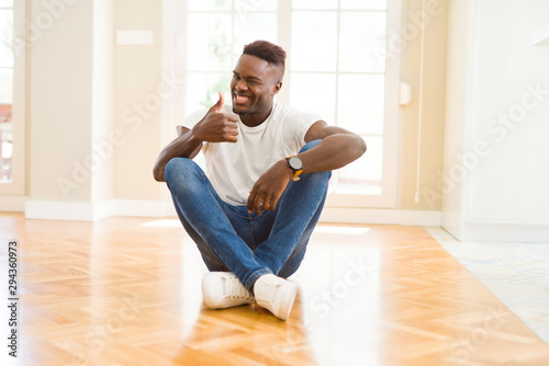 Handsome african american man sitting on the floor at home doing happy thumbs up gesture with hand. Approving expression looking at the camera with showing success. © Krakenimages.com