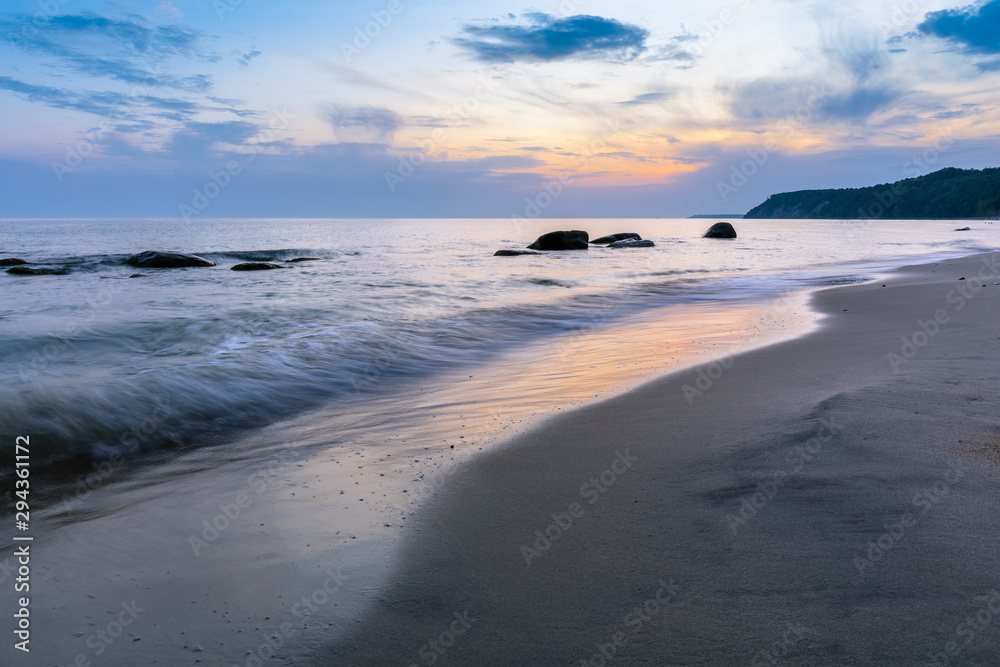 Obraz premium beautiful landscape of a wild sandy beach, a sea with protruding boulders on a long exposure and a steep coast on the horizon during sunrise