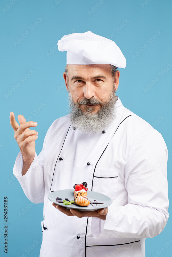 Waist up portrait of charismatic bearded chef presenting beautiful dessert while standing against blue background Waist up portrait of charismatic bearded chef presenting beautiful dessert while standing against blue background