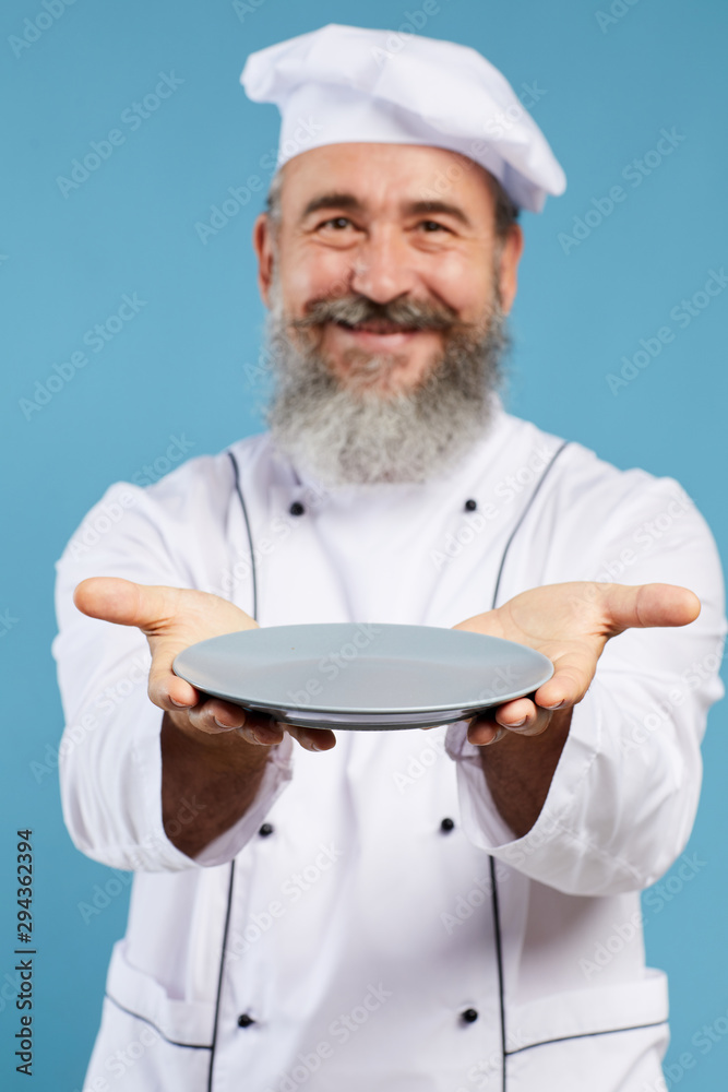 Portrait of bearded senior chef holding empty plate and smiling at camera while standing against blue background, mock up template, focus on foreground Portrait of bearded senior chef holding empty plate and smiling at camera while standing against blue background, mock up template, focus on foreground