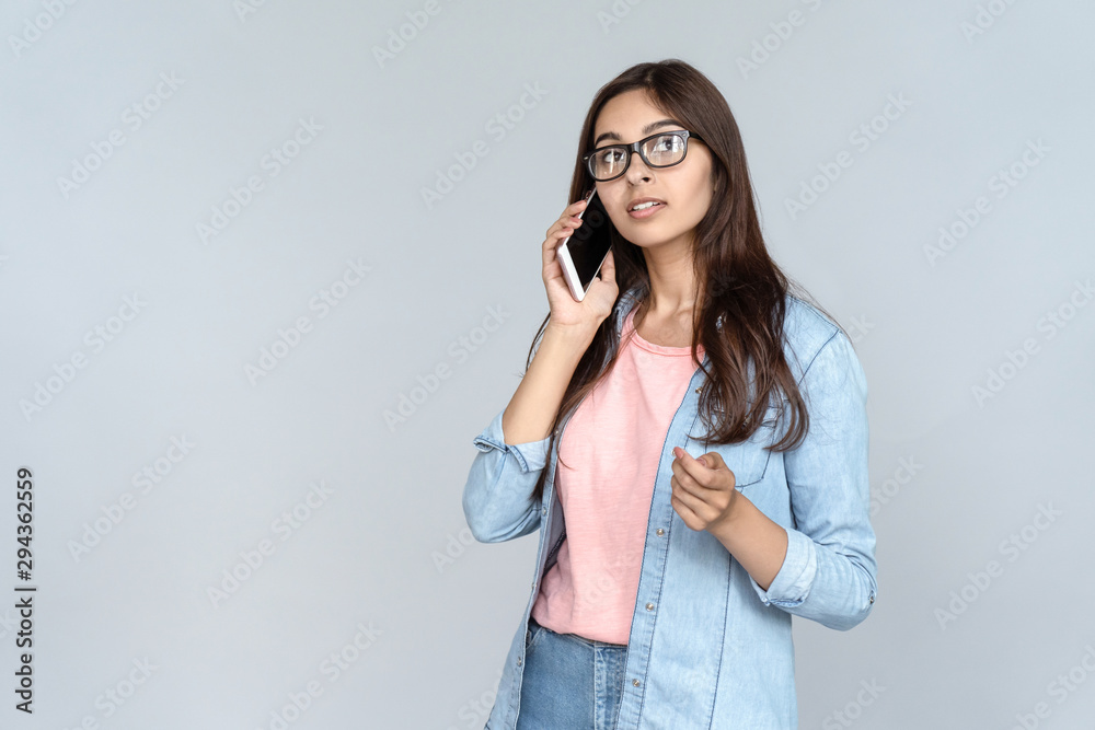 Serious young adult Indian woman student professional looking away talking on modern phone having mobile conversation speaking by cellphone standing isolated on grey background with copy space Serious young adult Indian woman student professional looking away talking on modern phone having mobile conversation speaking by cellphone standing isolated on grey background with copy space