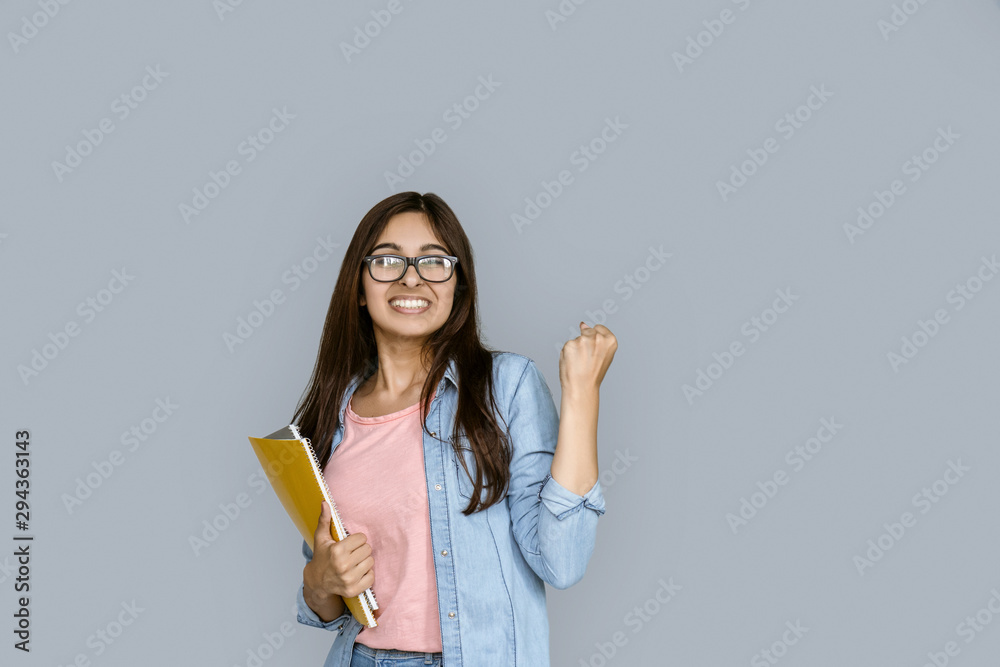 Happy young adult indian woman winner celebrate university admission scholarship good test exam result. Student girl hold copybook looking at camera standing isolated on grey background copy space Happy young adult indian woman winner celebrate university admission scholarship good test exam result. Student girl hold copybook looking at camera standing isolated on grey background copy space