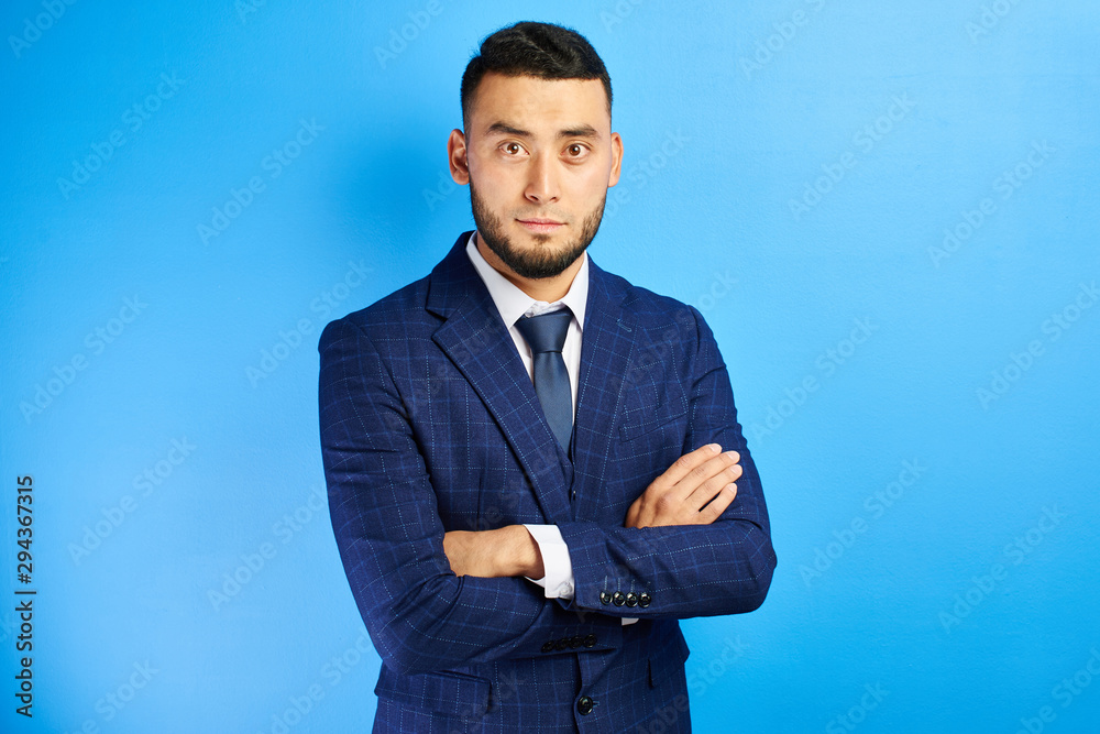 Portrait of serious handsome successful kazakh asian businessman in suit with arms crossed on blue studio background , isolated with copy space Portrait of serious handsome successful kazakh asian businessman in suit with arms crossed on blue studio background , isolated with copy space