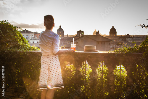 Canvas Print Young woman tourist fashion white dress with spritz cocktail in front of panoramic view of Rome cityscape from campidoglio terrace at sunset