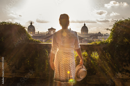 Canvas Print Young woman tourist in fashion white dress with hat at panoramic view of Rome cityscape from campidoglio terrace at sunset