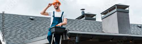 panoramic shot of happy repairman sitting on roof and holding toolbox photo