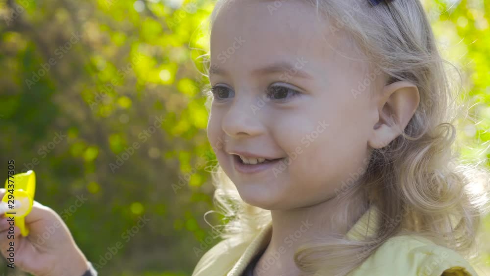 Side view of a cute blond girl blowing soap bubbles and smiling. Little charming child with brown eyes playing in the park in sunrays. Side view of a cute blond girl blowing soap bubbles and smiling. Little charming child with brown eyes playing in the park in sunrays.