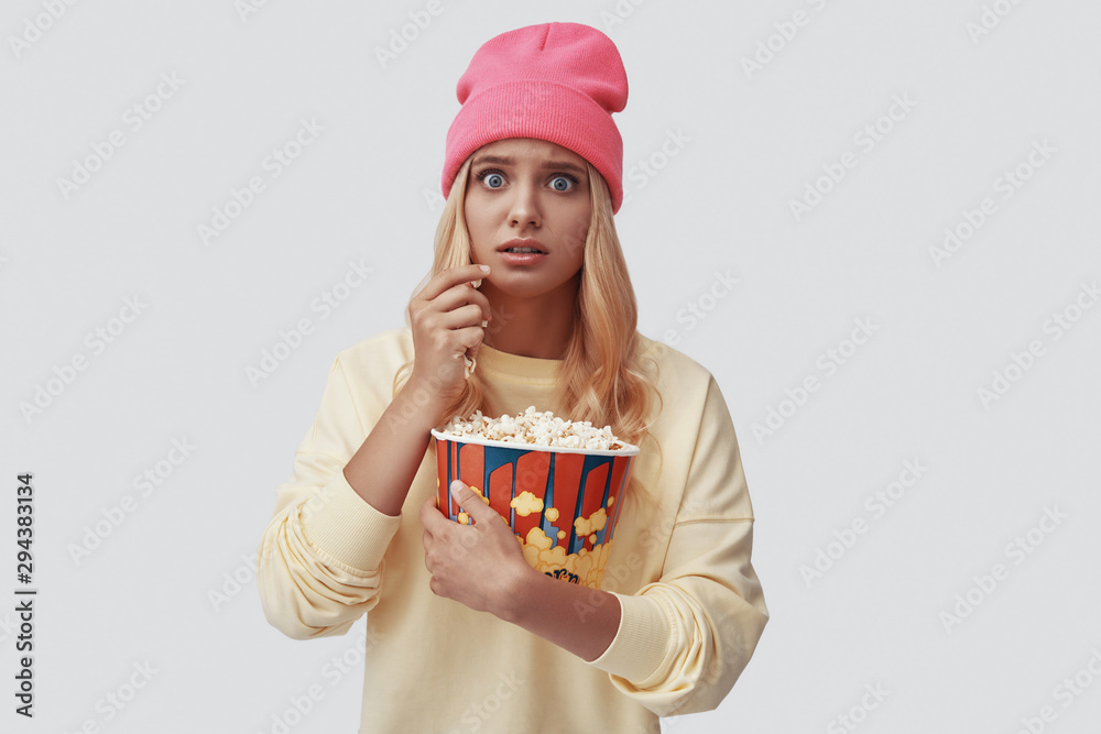 Attractive young woman eating popcorn and looking at camera while standing against grey background Attractive young woman eating popcorn and looking at camera while standing against grey background