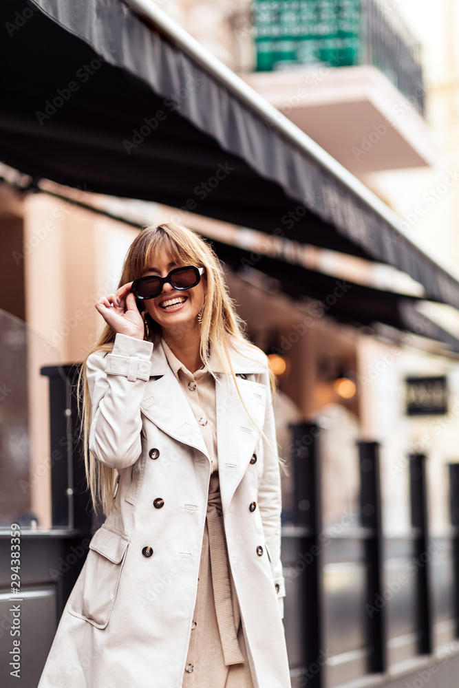 Stylish beautiful woman blonde in beige trench coat and brown sunglasses walking around the city