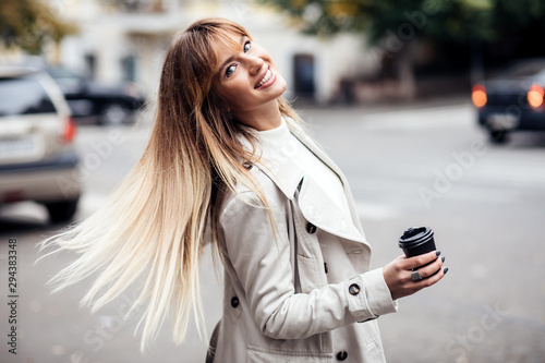 Stylish beautiful woman blonde in beige trench coat.She holds coffee to go. portrait of smiling girl . Street fashion concept