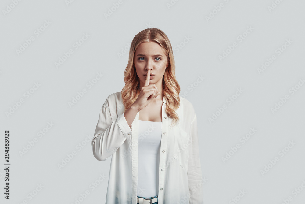 Beautiful young woman looking at camera and keeping finger on lips while standing against grey background Beautiful young woman looking at camera and keeping finger on lips while standing against grey background