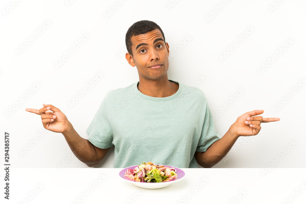 Young handsome man with salad in a table pointing to the laterals having doubts Young handsome man with salad in a table pointing to the laterals having doubts