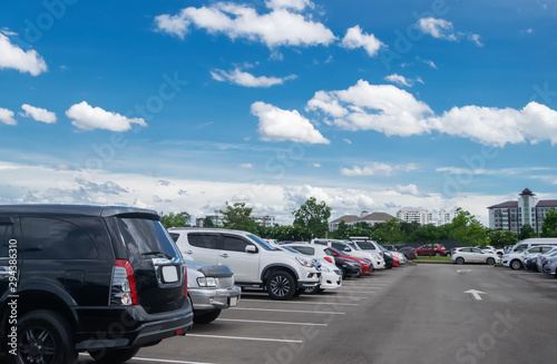 Car parking in large asphalt parking lot with trees, white cloud and blue sky background