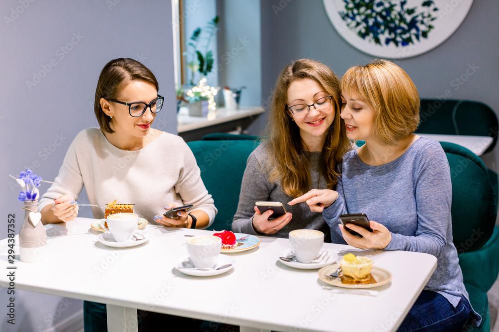 Three young women eating cakes and drinking coffee or tea at cafe, coffee break. Pretty girls sitting at cafe and using smartphones, making photos