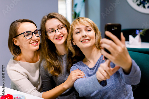 Three beautiful young women enjoying coffee and cake together in a cafe sitting at a table laughing and making selfie photo with happy smiles