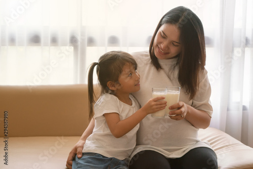 Little asian girl and her pregnant mom drinking milk sitting on sofa near the window at home. Motherhood and care, healthy eating and lifestyle.