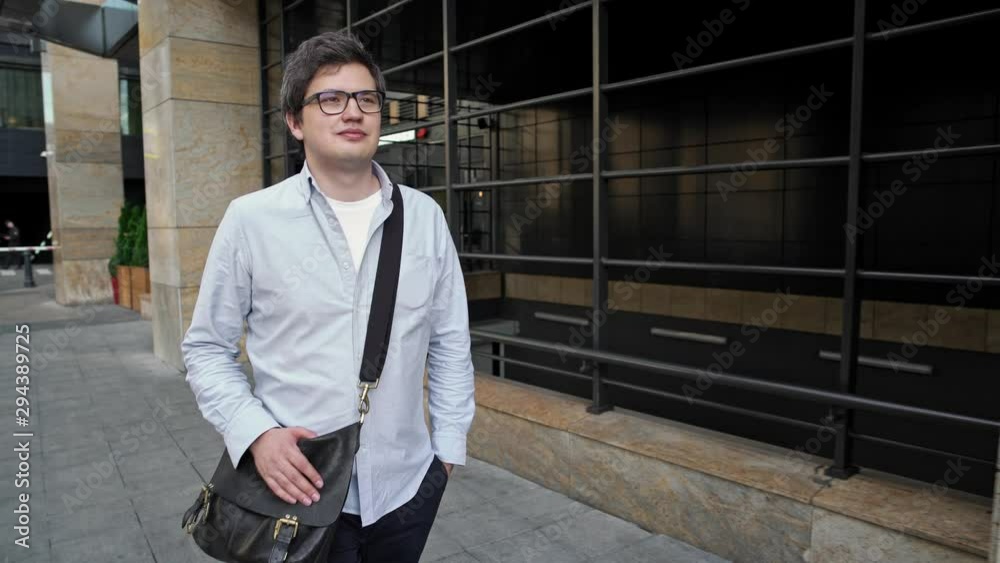 Middle shot of smiling man wearing blue shirt and shoulder bag in eyeglasses walking along the street on background of business center in daylight Middle shot of smiling man wearing blue shirt and shoulder bag in eyeglasses walking along the street on background of business center in daylight