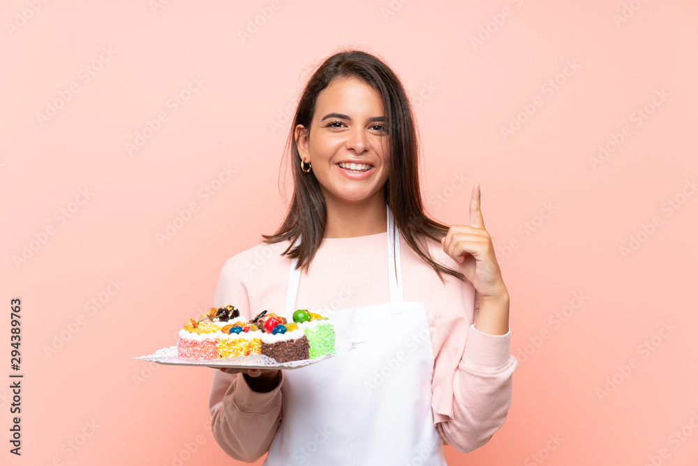 Young girl holding lots of different mini cakes over isolated background pointing up a great idea Young girl holding lots of different mini cakes over isolated background pointing up a great idea