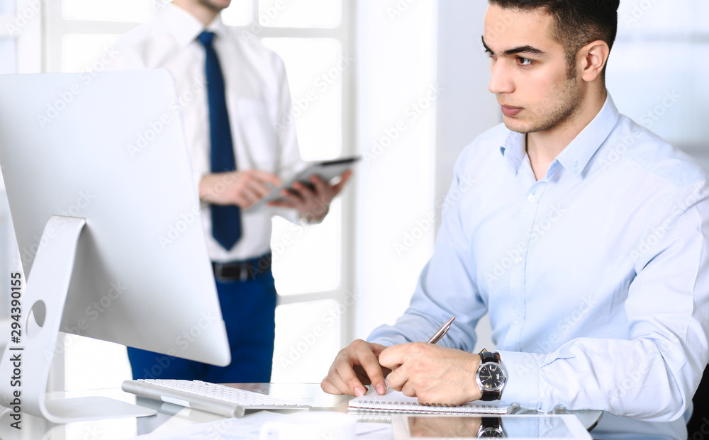 Obraz premium Businessman working with computer in modern office, male colleague at background . Headshot of arab male entrepreneur or manager at workplace. Business concept, teamwork and multi ethnic