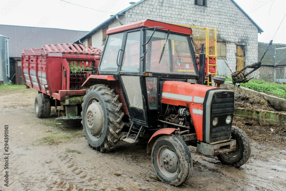 Red tractor with a trailer in the yard of a dairy farm. Side view. A barn in the background. Podlasie, Poland. Red tractor with a trailer in the yard of a dairy farm. Side view. A barn in the background. Podlasie, Poland.