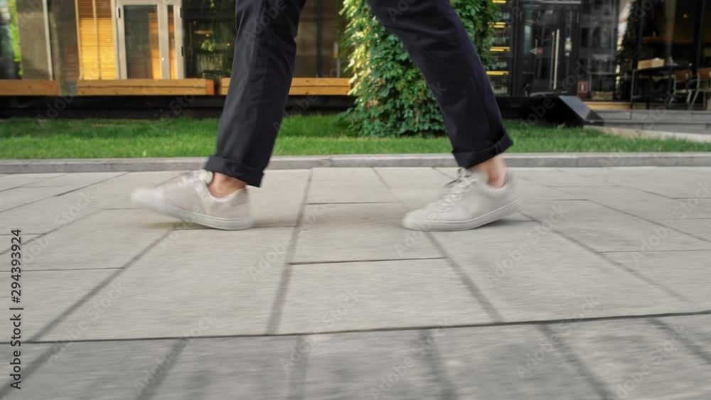Gimbal shot profile view from ground level of men wearing blue trousers and white sneakers walking along grey pavement on background of grass with sunlight Gimbal shot profile view from ground level of men wearing blue trousers and white sneakers walking along grey pavement on background of grass with sunlight