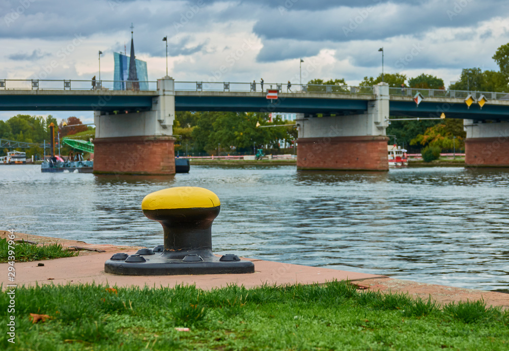 Fototapeta premium Yellow bollard for the line for mooring riverboats, with river and bridge blurred in the background (selective focus on the bollard)