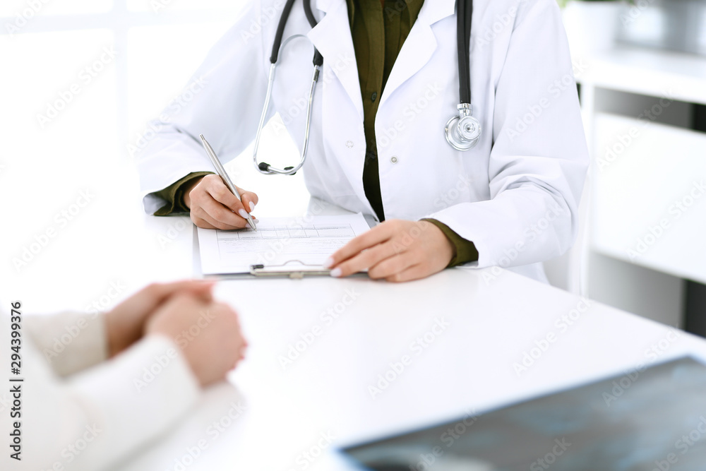 Woman doctor and patient sitting and talking at medical examination at hospital office, close-up. Therapist filling up medication history records. Medicine and healthcare concept Woman doctor and patient sitting and talking at medical examination at hospital office, close-up. Therapist filling up medication history records. Medicine and healthcare concept